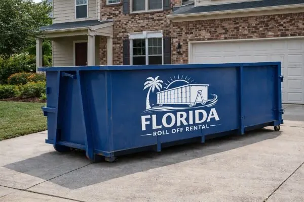 Blue roll-off dumpster parked in the driveway of a brick two-story house