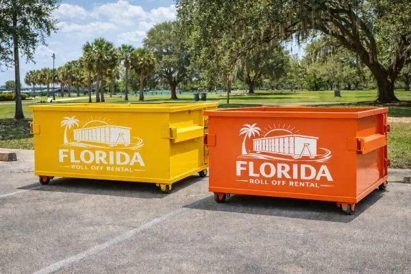 Yellow and orange dumpsters side by side in a paved parking lot with palms and live oaks behind